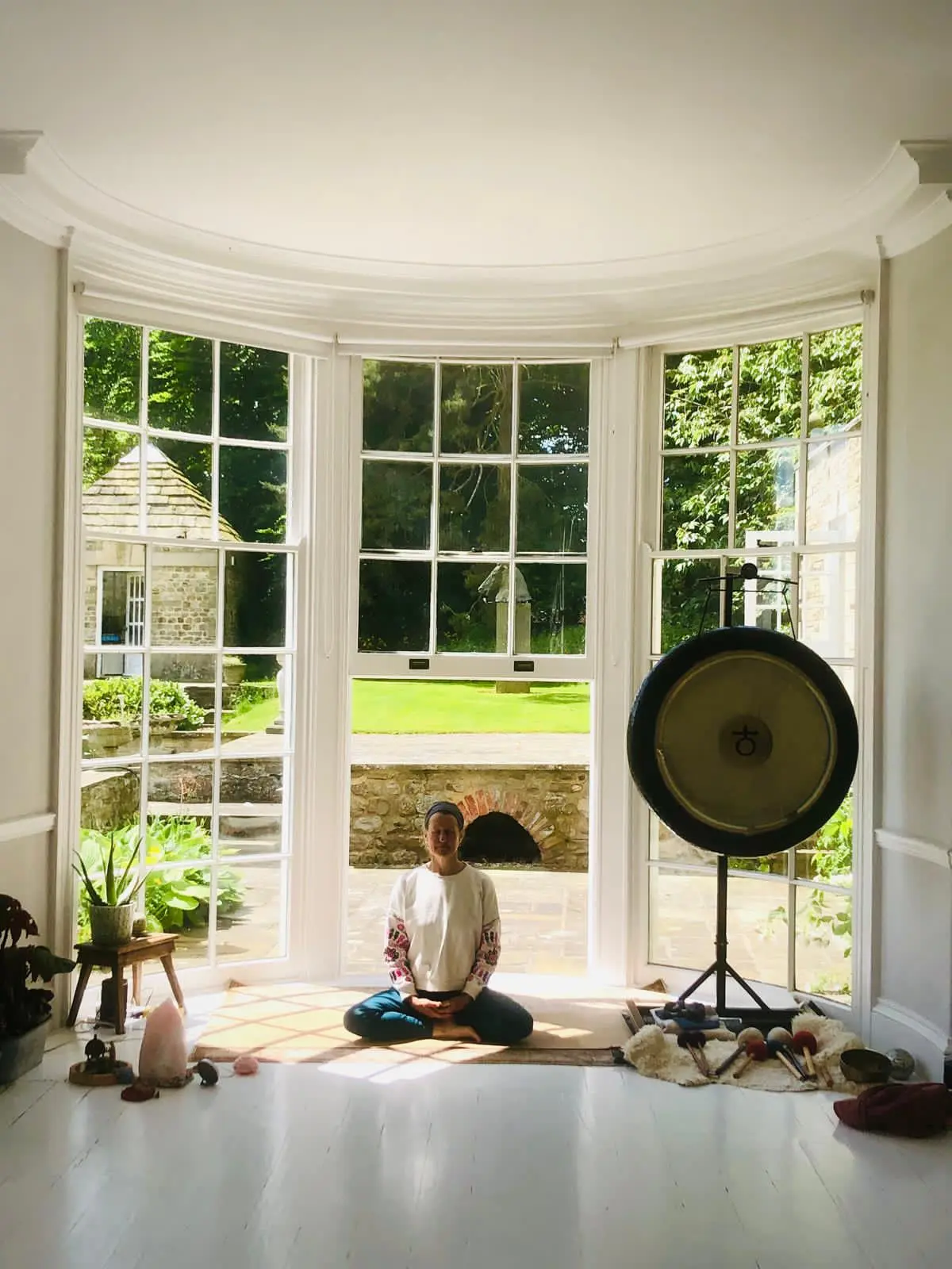 Rose Long sat leading a retreat class in cross-legged position on the floor, smiling with her eyes closed. She is sat in-front of a large bay window which looks out onto a large sun-filled garden. There is a large, golden gong to her left and a plant with a himilayan salt lamp on her right.
